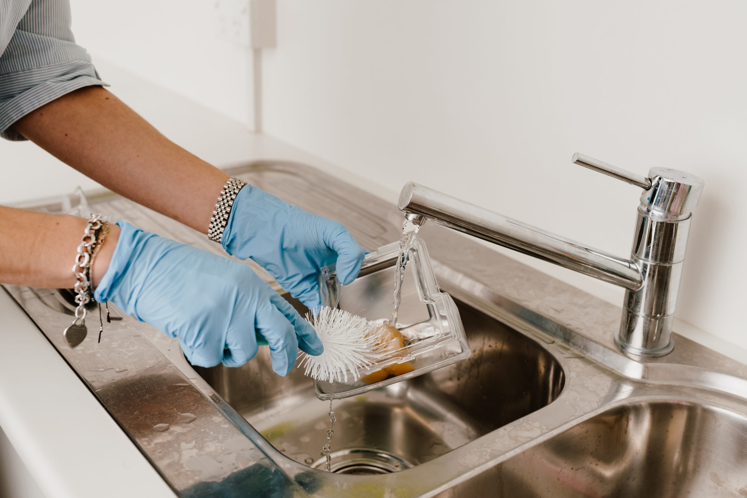 Gloved hands rinsing a humidifier chamber under a running tap, cleaning inside it with a white bottle brush