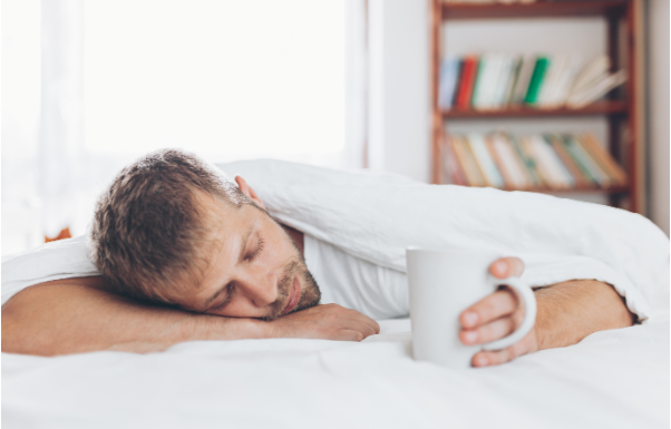 A man asleep face-down in bed, one hand still holding a mug of coffee